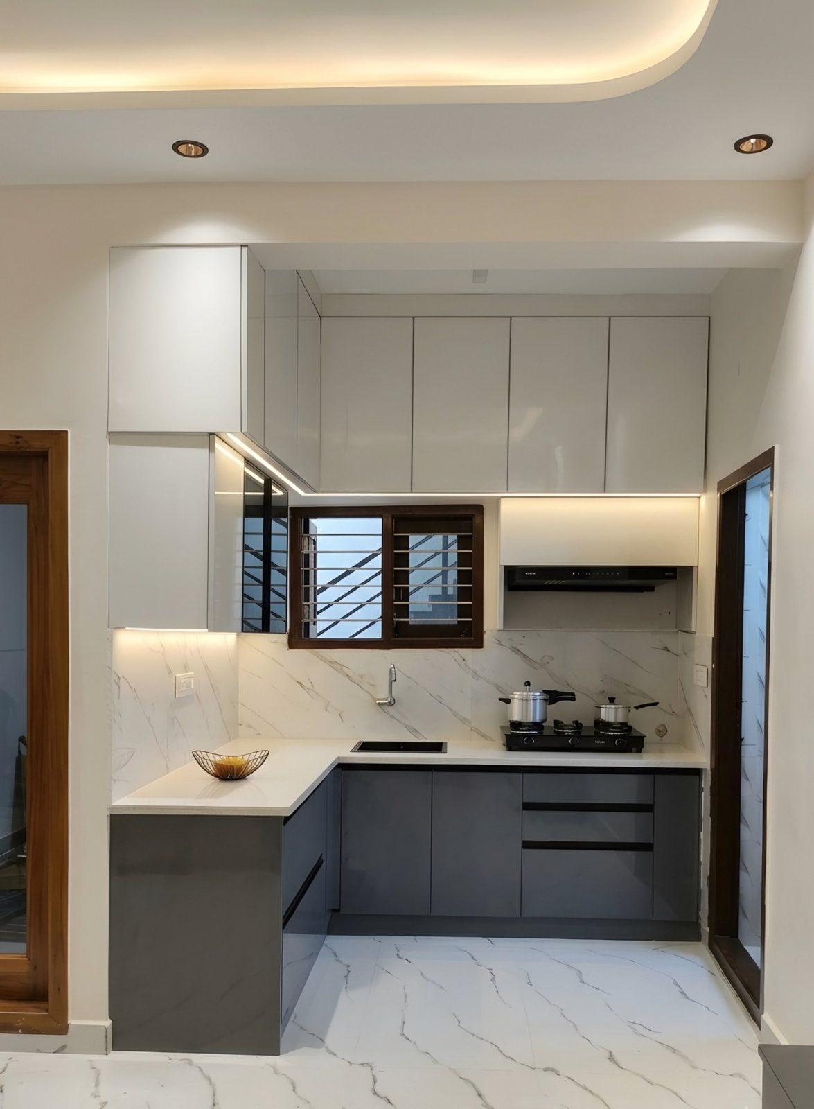 L-shaped modular kitchen in a Hassan 2 BHK with grey base units, white gloss overheads, marble backsplash and profile lighting