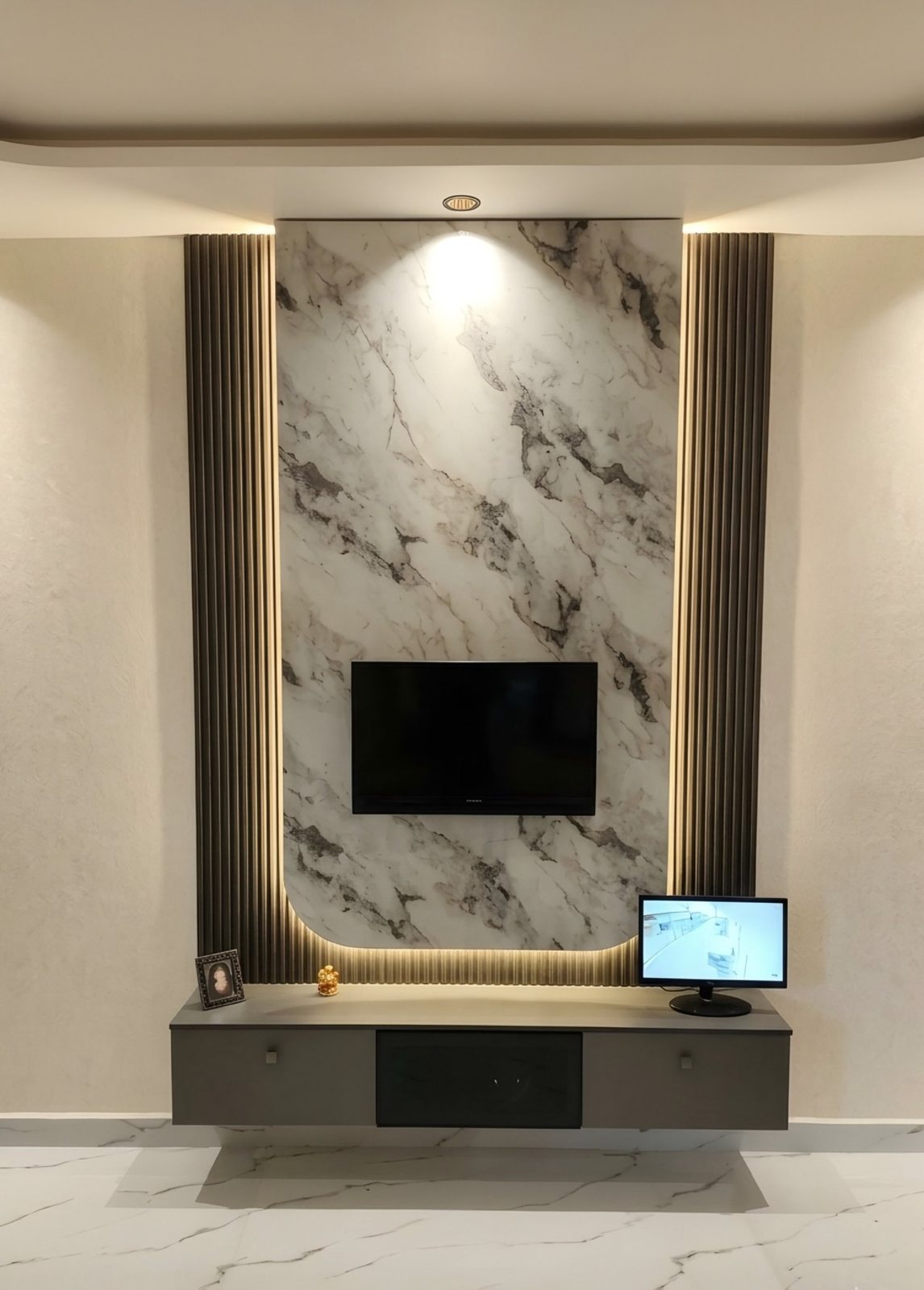 Living room TV unit design in a 2 BHK in Hassan, Karnataka — backlit marble-finish panel with fluted side panels and floating grey console