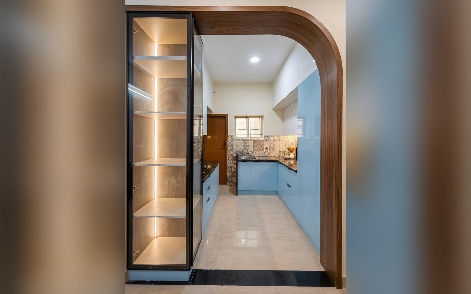 Sky-blue modular kitchen seen through a curved walnut archway with a backlit glass display unit, Surya Elegance, Chandapura